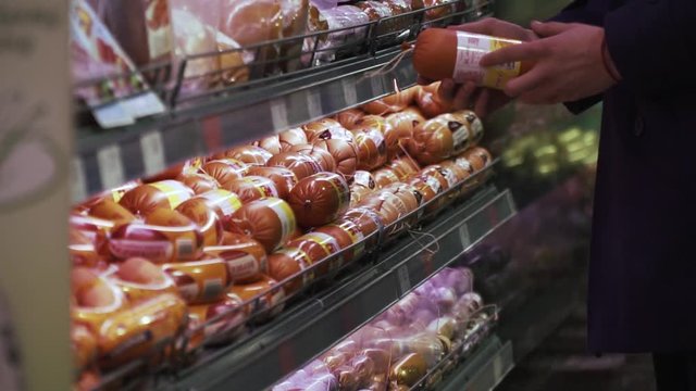 A Man Is Picking Sausages In The Supermarket, Long Aisle Full Of Goods