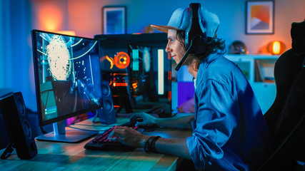 Professional Gamer Playing First-Person Shooter Online Video Game on His Powerful Personal Computer. Room and PC have Colorful Neon Led Lights. Young Man is Wearing a Cap. Cozy Evening at Home.