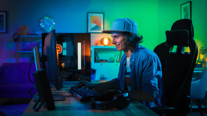 Professional Gamer Playing First-Person Shooter Online Video Game on His Powerful Personal Computer with Colorful Neon Led Lights. Young Man is Wearing a Cap. Living Room Lit with Green Lamps. Evening © Gorodenkoff