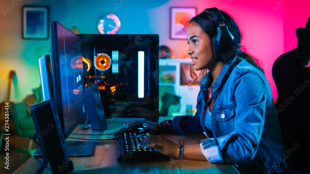 Wall mural Portrait of a Pretty and Excited Black Gamer Girl with Headphones Smiling to the Camera. She is Sitting on Chair next to Powerful Personal Computer. Home and PC is Lit with Neon Lights.