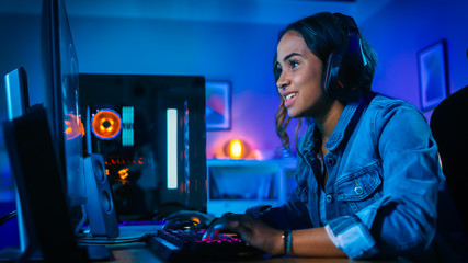 Pretty and Excited Black Gamer Girl in Headphones is Playing First-Person Shooter Online Video Game on Her Computer. Room and PC have Colorful Neon Led Lights. Cozy Evening at Home.