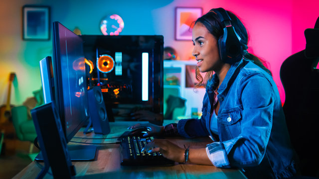 Portrait Of A Pretty And Excited Black Gamer Girl With Headphones Smiling To The Camera. She Is Sitting On Chair Next To Powerful Personal Computer. Home And PC Is Lit With Neon Lights.