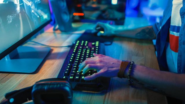 Close-Up Hands Shot Showing a Gamer Using the Keyboard while Playing an Online Shooter Video Game. Keyboard has Green Neon Lights in Buttons. Gamer is Wearing a Bracelet. Room is Dark.