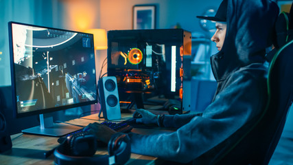 Professional Gamer and Streamer Playing First-Person Shooter Online Video Game on His Cool Personal Computer. Young Man is Wearing a Cap and Hood. Room and PC have Colorful Neon Led Lights.