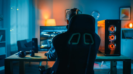 Back Shot of a Gamer Playing First-Person Shooter Online Video Game on His Powerful Personal Computer. Room and PC have Colorful Neon Led Lights. Cozy Evening at Home. - obrazy, fototapety, plakaty