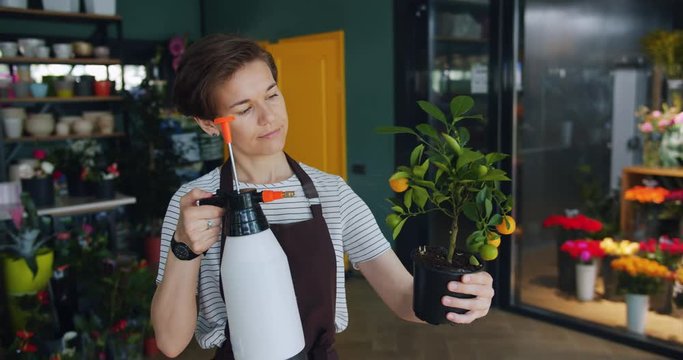 Cheerful young florist is watering cirtus plant in flower store using sprayer bottle enjoying job working with greenery. People and small business concept.