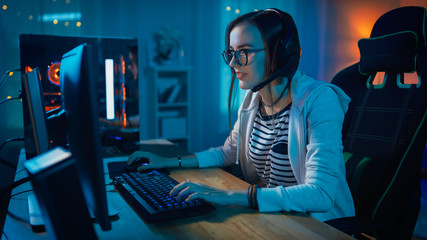 Excited Gamer Girl in Headset with a Mic Playing Online Video Game on Her Personal Computer. She Talks to Other Players. Room and PC have Colorful Warm Neon Led Lights. Cozy Evening at Home. - obrazy, fototapety, plakaty