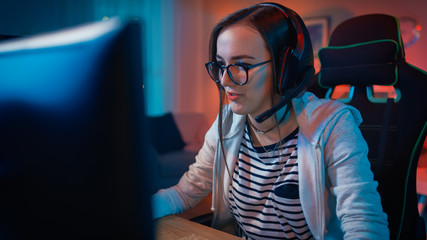 Excited Gamer Girl in Headset with a Mic Playing Online Video Game on Her Personal Computer. She Talks to Other Players. Room and PC have Colorful Warm Neon Led Lights. Cozy Evening at Home.