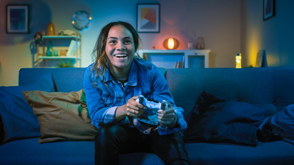 Beautiful Excited Young Black Gamer Girl Sitting on a Couch and Playing Video Games on a Console. She Plays with a Wireless Controller. Cozy Room is Lit with Warm and Neon Light. © Gorodenkoff