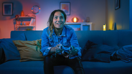 Beautiful Excited Young Black Gamer Girl Sitting on a Couch and Playing Video Games on a Console. She Plays with a Wireless Controller. Cozy Room is Lit with Warm and Neon Light. © Gorodenkoff