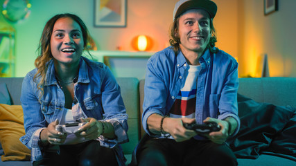 Excited Black Gamer Girl and Young Man Sitting on a Couch and Playing Video Games on Console. They Plays with Wireless Controllers. Cozy Room is Lit with Warm and Neon Light.