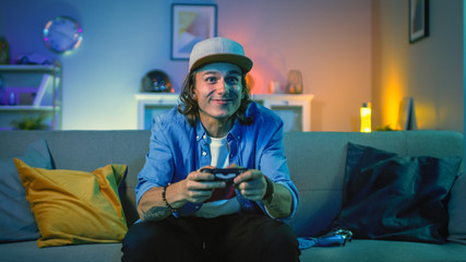 Handsome Excited Young Gamer with Long Hair and a Cap is Sitting on a Couch and Playing Video Games on a Console. He Plays with a Wireless Controller. Cozy Room is Lit with Warm and Neon Light. © Gorodenkoff