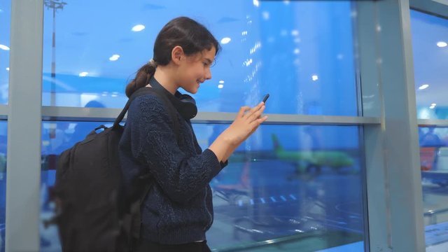 daughter with a backpack airplane flights airport concept. girl teenager with a smartphone lifestyle looks out the window at the airport area airplanes and bus at a distance. teen girl looks at the