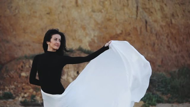 pretty happy girl in black gymnastic swimsuit poses with piece of white cloth on hips on sea beach by hill slow motion