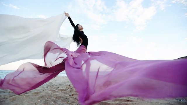 joyful barefoot woman in bodysuit with large transparent cloth sheets stands on beach sand in strong wind slow motion