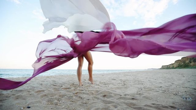 brunette lady with slim legs poses holding large chiffon pieces waved by wind on empty coast under blue sky slow motion. Classic dancer on the nature