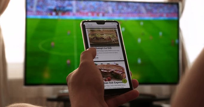 Against the background of the TV at home (at the bar), the guy (woman) orders different snacks, pizza and sweets scrolling through internet stores using mobile phones. Concept: Football, Sport,Order.