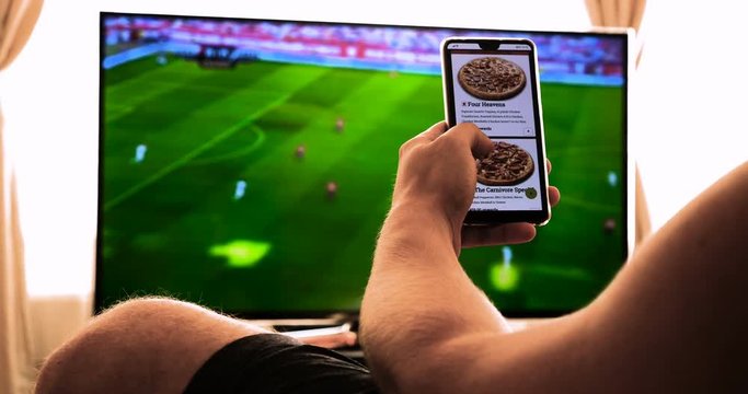 Against the background of the TV at home (at the bar), the guy (woman) orders different snacks, pizza and sweets scrolling through internet stores using mobile phones. Concept: Football, Sport,Order.
