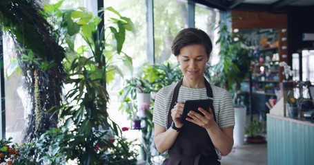 Pretty female florist is counting plants entering records in tablet busy with work working in shop walking around smiling. People, business and job concept.