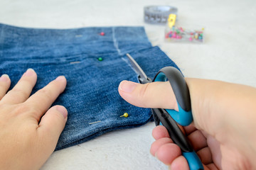 Woman hand holding a scissors and cutting a blue jeans shorts to make them shorter. Making denim shorts. On a white.