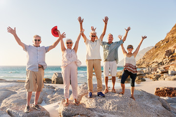 Portrait Of Senior Friends Standing On Rocks By Sea On Summer Group Vacation With Arms Outstretched