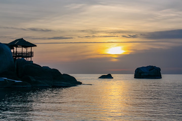 Naklejka na ściany i meble Beautiful sunset on the paradise island of Thailand. Tropical bungalow on a mountain with stones against the background of the sea and the sun