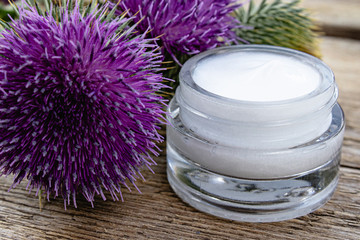 Obraz na płótnie Canvas Thistle flower with open jar of cosmetic face cream on wooden background. Medicinal plant.