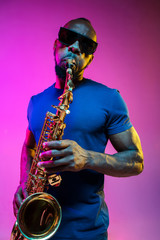 Obraz na płótnie Canvas Young african-american jazz musician playing the saxophone on pink studio background in trendy neon light. Concept of music, hobby. Joyful attractive guy improvising. Colorful portrait of artist.