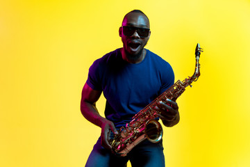 Fototapeta na wymiar Young african-american jazz musician playing the saxophone on yellow studio background in trendy neon light. Concept of music, hobby. Joyful attractive guy improvising. Colorful portrait of artist.