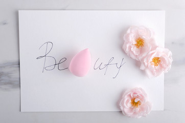 Top view of written wird Beauty, pink cosmetic sponge and roses on the marble surface