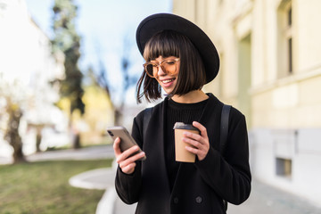 Young woman in black hat standing at the street drinking coffee to go and using mobile phone
