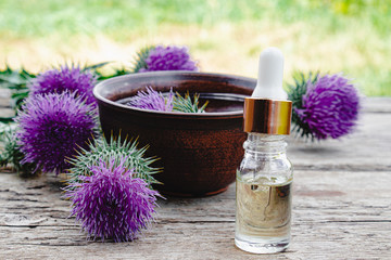Thistle essential oil in a bottle on the table near the thistle flowers on wooden background.