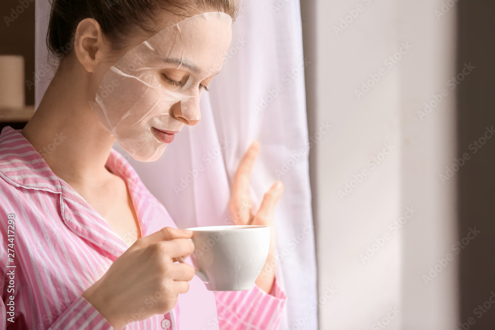 Wall mural young woman with sheet facial mask drinking tea at home