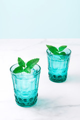 Two beautiful vintage turquoise glasses with cold drink and ice cubes, decorated with fresh green mint leaves on white marble table. Close-up.