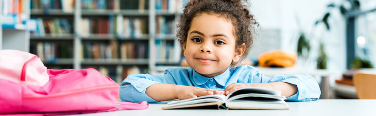 panoramic shot of happy african american kid sitting near book in library