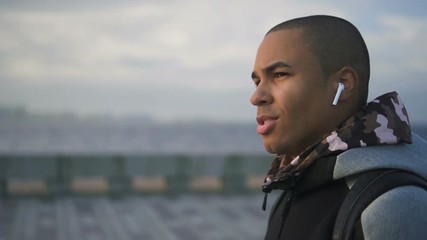 Portrait of african american man in stylish earphones walking on urban embankment. Ethnic male talking to bass about career using digital devices and smiling on riverside.