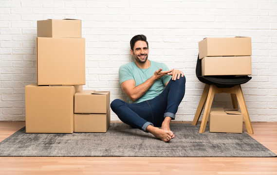 Handsome young man moving in new home among boxes extending hands to the side for inviting to come