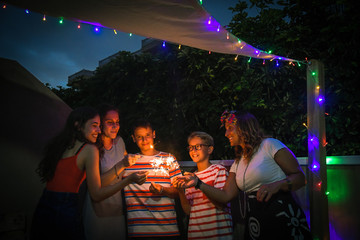 Group of various people making party with fireworks outdoor in terrace. Smiling girls and boys having fun together enjoying sparklers celebrating family birthday. Youth happiness and holiday concept