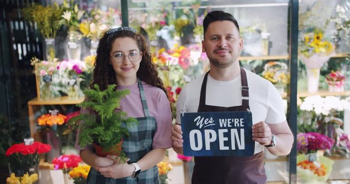 Couple of florists holding open slate and green plant standing together in flower shop smiling looking at camera with colorful beautiful bouquets in background.