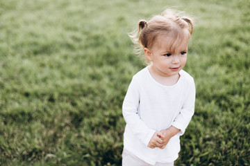 little baby girl with two tails, wearing a white T-shirt standing on green grass in nature, smiling. Child in the park looking very happy