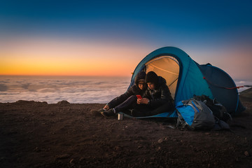 Two boys playing with smartphone sitting in a camping tent. Couple of young tourists enjoying high mountain vacation at sunset. Beautiful panorama in background. Adventure trip and open air concept