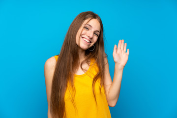 Young woman with long hair over isolated blue wall saluting with hand with happy expression © luismolinero