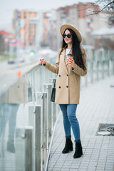 Young woman wearing coat and hat walking in the autumn city street and drinking take away coffee in...