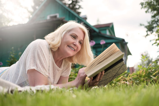 Caucasian mature blonde woman reading a book in the garden. Moment of leisure on summer day. House is on background.