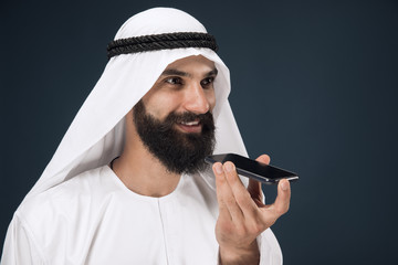 Half-length portrait of arabian saudi man on dark blue studio background. Young male model using smartphone, making a call. Concept of business, finance, facial expression, human emotions