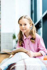 selective focus of cute and cheerful kid sitting and holding book in library