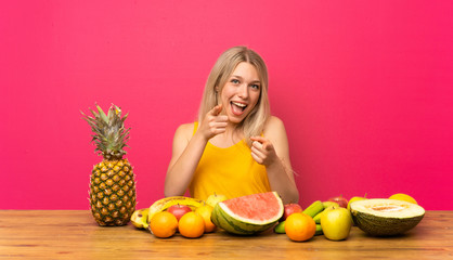 Young blonde woman with lots of fruits points finger at you