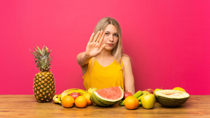 Young blonde woman with lots of fruits making stop gesture with her hand