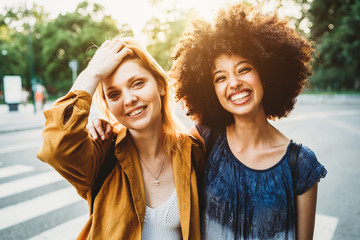 Porträt von zwei Freundinnen bei Sonnenuntergang - Zwei Millennials, eine Blondine und ein schwarzes Mädchen mit Afrohaar schauen in die Kamera und umarmen sich