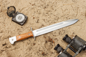 Hunter knife with wooden handle lies on the sand with old military binoculars and old compass....